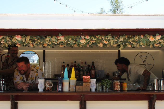Two bartenders prepare drinks at a stylish outdoor bar with floral decor.