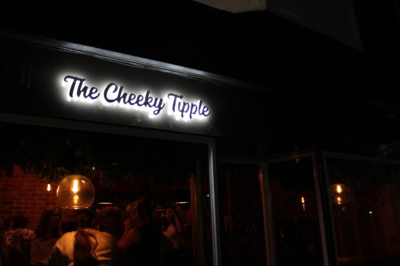 Sign for "The Cheeky Trip" illuminated at night, with a warm, inviting atmosphere.